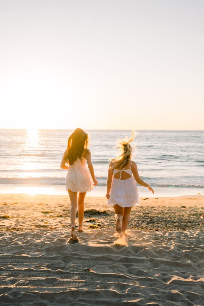 Friends in white dresses running towards the beach at sunset during a Los Angeles senior photo session.