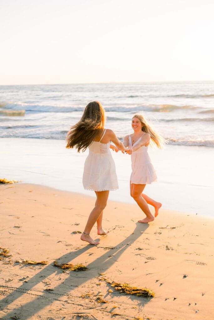 Best friends in white dresses twirling on the beach at sunset.