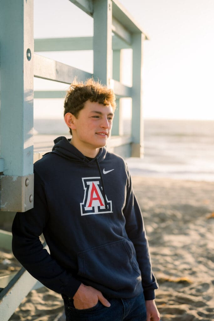 A senior boy in a college sweatshirt posing on the beach for his senior photos.