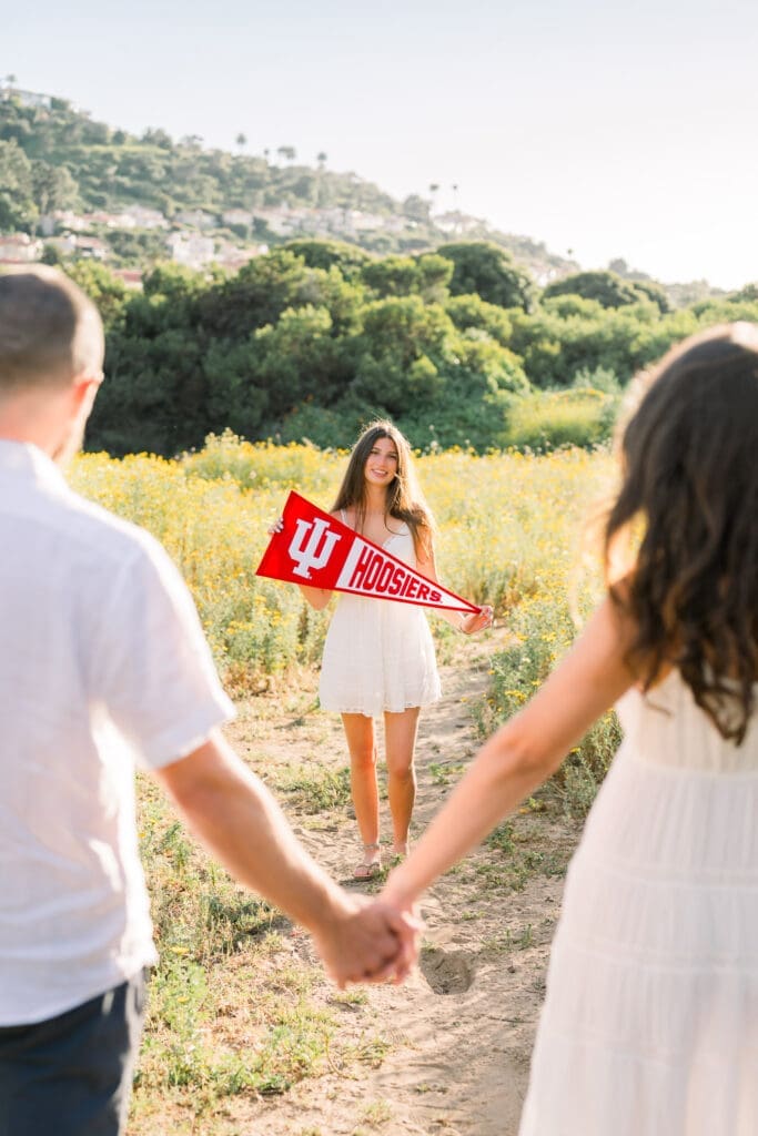 Mom and dad hold hands while looking at their senior daughter holding her college banner.
