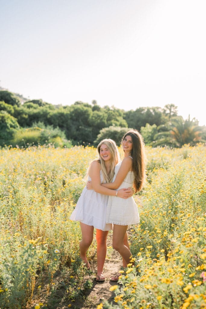Best friends in white dresses laughing in a field of wildflowers while posing for their Los Angeles senior photo session.