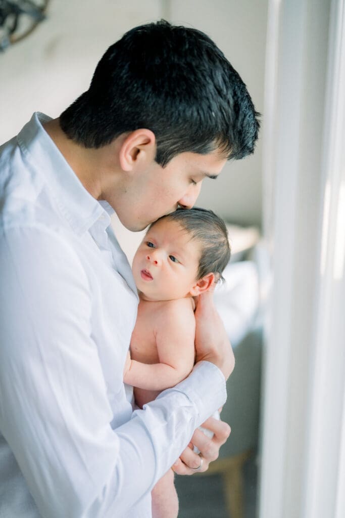 A dad kissing his newborn son's head while taking newborn photos at home.