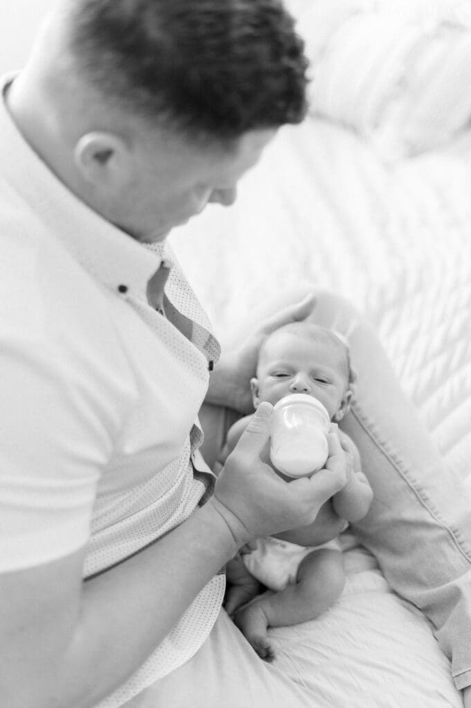 A dad feeding his newborn baby a bottle while taking newborn photos at home.