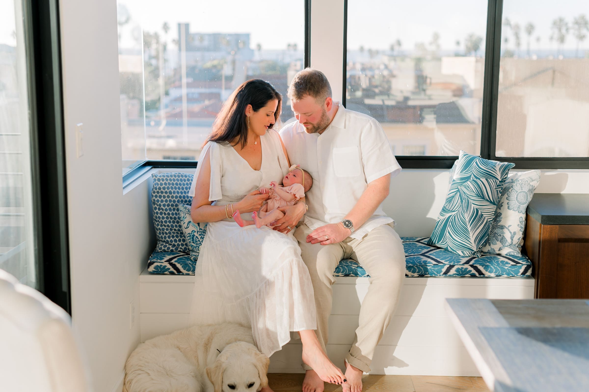 A guide for what to wear for newborn photos at home featuring a mom and dad in light natural colored clothing holding their newborn baby in a pink dress.