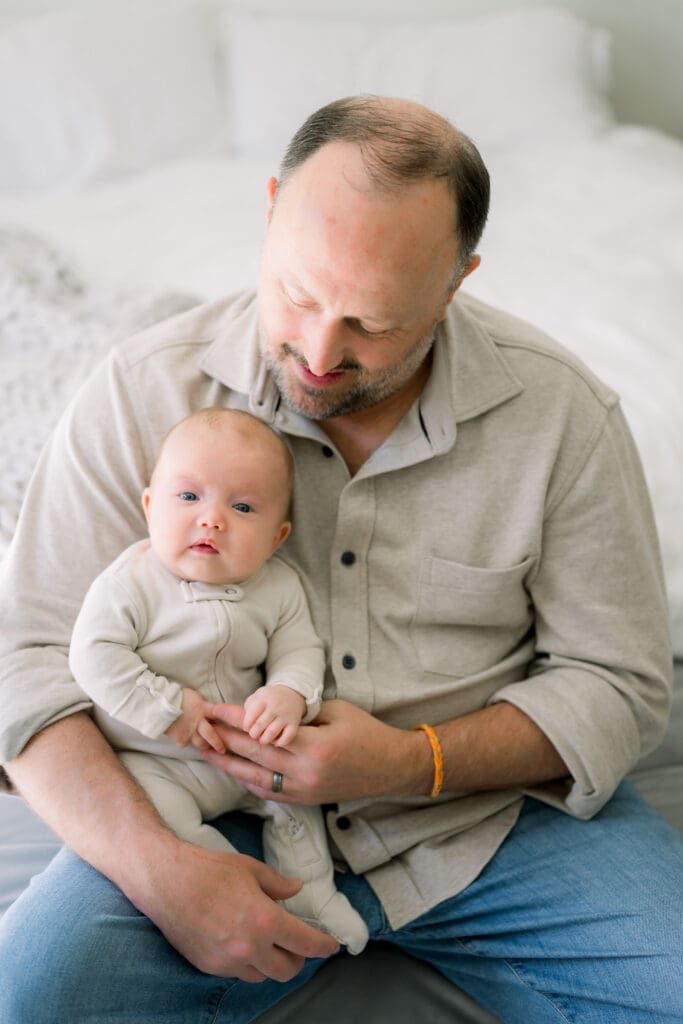 A dad holding his newborn baby at home.  They are wearing tan colored clothing that is perfect for what to wear for newborn photos.