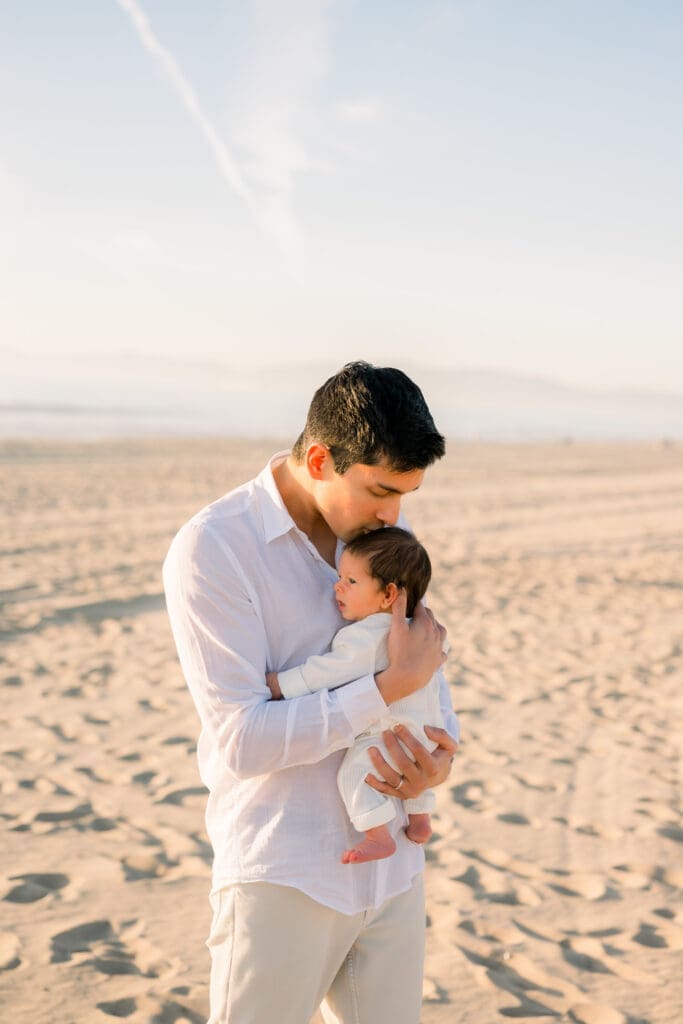 A dad kisses the top of his son's head during an outdoor newborn session on the beach.