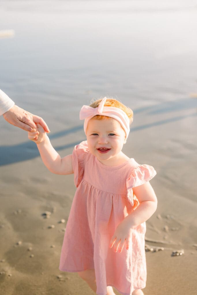 A little girl in a pink dress holding her mom's hand on a beach.
