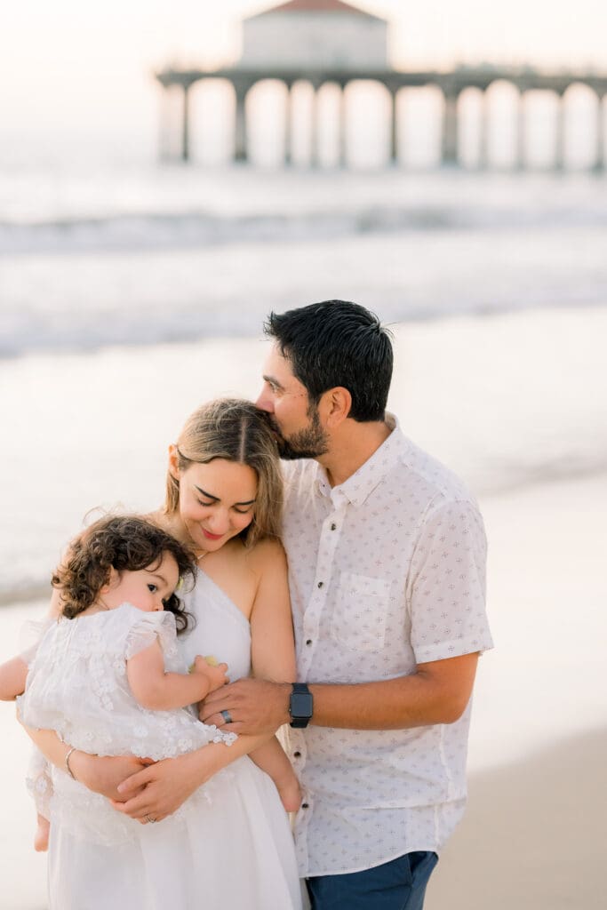 A husband kissing his wife's head while they hold their little girl at the beach.